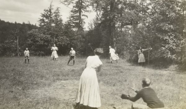 A baseball game is underway on the Island. A woman, with her back to the camera, is at bat. From left to right on the field: Donald Holt, Jeannette Holt, Alfred Holt, a woman running to second base, possibly Anna Holt Wheeler with Lillian Wheeler, and possibly Eleanor Holt with her arm on the post. The catcher has his back to the camera. 

