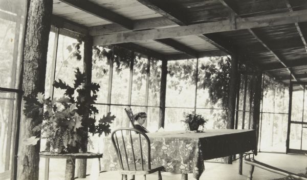 Seen in profile, Jeannette Holt is sitting in a rocking chair and reading a book, on the new porch addition to Island Lodge. A table with tablecloth and chair are in the center of the porch. A fern in a pot is sitting on the table. Caption reads: "Porch Addition."