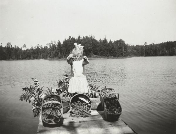 View from shoreline towards a girl, possibly Lillian Wheeler, standing on the pier at the Ark cottage on a sunny day. She is holding up some fruit and is wearing a dress, a bow in her hair. In front of her are three propped up wooden baskets filled with more fruit. There are two more wooden baskets filled with fruit next to her, and behind those baskets is some foliage. In the background is Archibald Lake and the wooded shoreline.