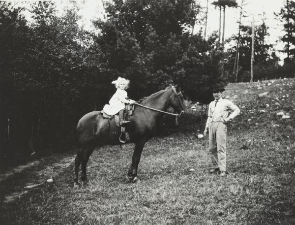 Looking at the camera, a girl, possibly Lillian Wheeler, is holding the reigns and sitting astride a horse. She is wearing a dress with pants underneath, a her boots are in the stirrups. The horse is standing in profile. A man, possibly Gus, wearing a cowboy hat, and shirt, pants and necktie, is standing in front of the horse looking towards the camera.
 