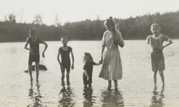 Lillian Wheeler is wading in Archibald Lake. She may be holding the hand of Jeannette Holt. There is another girl who is wearing bloomers, which may be Eleanor Holt, on the far right. The boys may be, from left to right, Alfred Holt and Donald Holt. All of the children are slim. 
