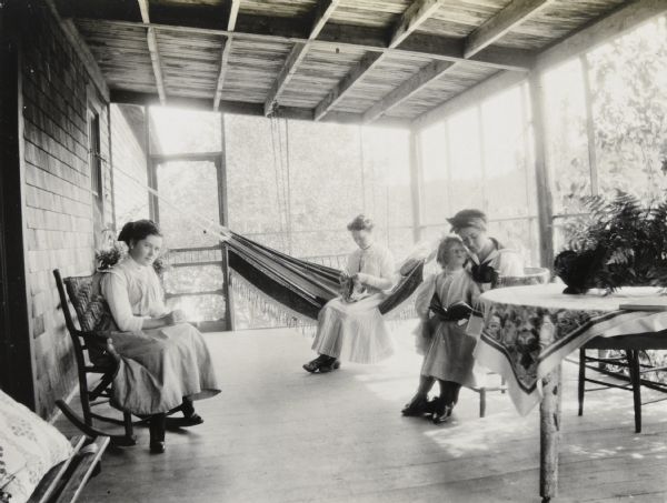 A small group is gathered on the porch of the Ark cottage in the daytime. Looking at the camera, a young woman, possibly Rose or Agnes Wilson, is sitting in a rocking chair. Sitting on the fringed hammock, Anna Holt Wheeler is doing some handicraft. By the table, Lillian Wheeler and a woman, possibly Rose or Agnes Wilson, are reading a book together. Caption reads: "Rose and Agnes Wilson at the Ark."