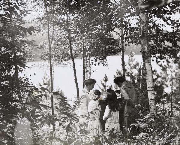 Two women and two girls are standing together under the trees and looking down into a large bucket. The girl on the left with a bow in her hair may be Jeannette Holt. Archibald Lake is in the background. 