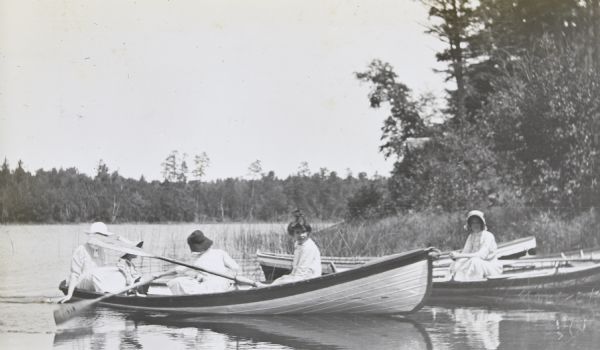 A group of four women are landing at Picnic Island in a clinker-built duck boat. One woman, sitting in the front of the boat, is looking at the camera. Behind the boat is another parked on the shoreline. The fifth woman, sitting in this boat with clasped hands, is also looking at the camera. Archibald Lake is in the background. 



