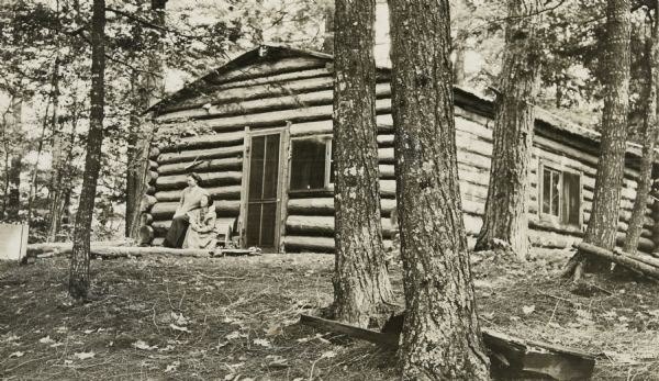 Two women are sitting on a bench outside of the Hermit cottage. The older woman has her arm around the neck and on the shoulder of the younger woman. They are both looking off in the distance. There are tree trunks in the foreground. Page heading reads: "The Hermit."

