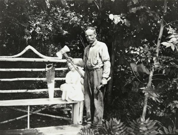 William Pettigrew, who was Foreman at Camp Four for sometime beginning in 1885, is holding a double headed ax in his right hand and a hammer in his left hand. There is a crosscut hand saw leaning against the wooden bench. The back of the bench is made with birch logs. Lillian Wheeler is perching in the corner of the bench. This location is probably near the Ark cottage. Trees and plants are in the background. Caption reads: "William Pettigrew."