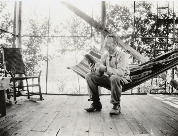 William Pettigrew, who was Foreman at Camp Four beginning in 1885, is reading a hard cover book on the porch of the Ark cottage while sitting on a striped hammock. He is wearing a striped shirt, pants, suspenders, and a tie tucked into his shirt. There is a woven cane backed rocking chair in the corner. In the background, visible through the porch screens, are trees. Caption reads: "Inside Screen House."