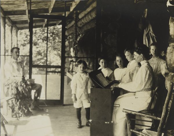 Lucy Rumsey Holt is playing the pump organ during Sunday church service on the Island Lodge porch. Her brother-in-law Grant Stroh is sitting in the corner by the screen door. A group of children are standing around the organ and singing. Names from left to right: Donald Holt, unknown, Eleanor Holt, Alfred Holt, and Jeannette Holt. Caption reads: "Church service." 

