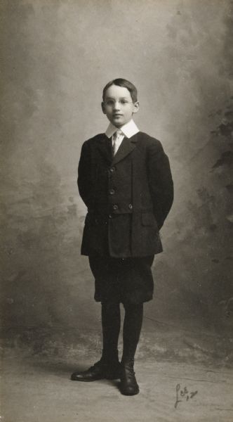 Full-length studio portrait of Donald Holt. He has his hands behind his back, and is wearing a coat, collared shirt, striped tie, short pants, stockings and shoes. The painted background is trees and mist.