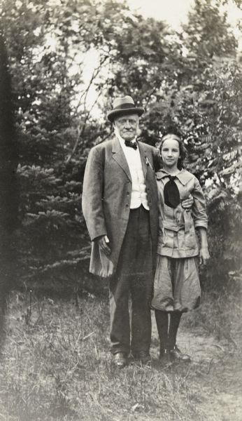 Grandfather I.P. Rumsey and granddaughter Eleanor Holt are standing together outdoors on the Island at Archibald Lake. I.P. Rumsey is wearing a suit, bow tie, and Homburg hat. Mary Eleanor Holt is wearing a shirt, tie, and knickers. Trees are in the background.