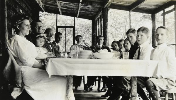 Lucy and W.A. Holt, with their family, the Trowbridge family and others, are sitting on the screened porch at Island Lodge for Sunday dinner. A tablecloth is over the wooden table, and a fern is in the center of the table. There is food in a bowl ready to be served. Trees are in the background. 

Names from left to right: Mrs. Trowbridge, Jane Trowbridge, Mr. Trowbridge, I.P. Rumsey, Calvin Trowbridge, Lucy Holt, W.A. Holt, Katherine Trowbridge, Alfred Holt, Cornelius Trowbridge, Donald Bridgeman, and Don Holt. Caption reads: left side: "Mrs. T, Mr. T., Jane, IPR, Calvin; right side: Katherine, Corneil, Donald Bridgeman, Don."