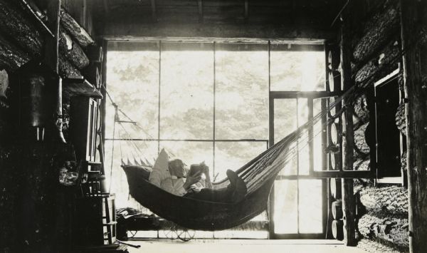 I.P. Rumsey is reading a book while lying in the hammock on the porch at Island Lodge. A pillow is tucked under his head. He is dressed in a long sleeved shirt, bow tie, and pants. On the left is a wooden chair, bookshelf, cabinet, and shelf. There is also a large metal urn with a spigot on the wall on the left.