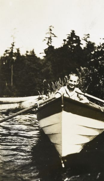 View towards Donald Holt sitting in a rowboat and smiling at the camera. He is on Archibald Lake with the shoreline just behind him.