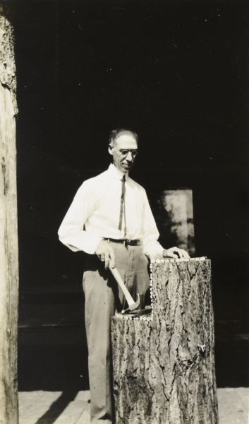 A man, holding a hammer, is standing behind the new log pulpit to be used for Sunday worship on the porch at Island Lodge. The base of the pulpit is a hollow hemlock log. The top surface of the pulpit is peeled birch branches laid side by side and nailed to the hemlock log. He is standing in sunlight, with the porch behind him in shadow. 