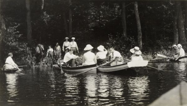 View across water towards a wooded shoreline, where a man and woman, followed by two children, are walking out of the woods toward Middle Lake where four wooden boats filled with people are tied up. Caption reads: "Middle Lake Start."