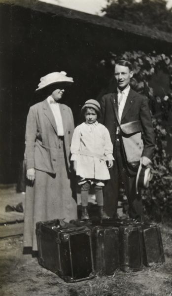 View of a child who is looking at the camera while standing on leather suitcases in front of Island Lodge on Archibald Lake. The child's mother and father, presumably, are standing behind the child on either side. The woman is wearing a long skirt, jacket, and shirt, and a large hat that is shading her face. The man is wearing a suit and tie, with the tie tucked in. He is holding a wide brimmed hat in his left hand and is wearing a cross body leather bag over his shoulder.