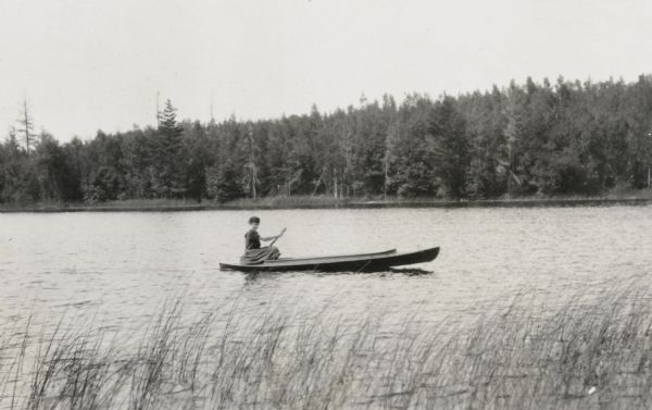 View from shoreline towards a girl paddling a boat on Archibald Lake. She is looking towards the camera. Trees are along the shoreline in the background.