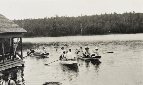 Elevated view from shoreline towards a group of nine people sitting in three rowboats on Archibald Lake near the boathouse on the left. Several of the boaters are smiling at the camera and waving. There is a boy peeking from behind a beam on the side of the boathouse. The opposite shoreline is in the background. Alfred or Donald Holt is in the left foreground.