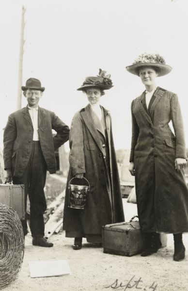From left to right: Albert Rhode, Mrs. Vendt and Minnie Vendt are standing with luggage. Mrs. Vendt is carrying a Swift's Silver Leaf Brand Lard tin pail. Minnie Vendt is wearing an overcoat and a hat decorated with artificial flowers. In the left foreground is a roll of wire and several squares of material.



