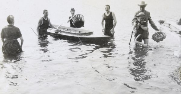 View towards a group digging for fresh water mussels in Archibald Lake. From left to right: a woman waist-deep in the water; a boy holding onto the side of a boat; a girl standing behind the boat with a large wooden basket; a boy smiling and wearing a tank-style bathing suit; Grant Stroh with his daughter Juliet Stroh, who has a tool in her hand. Caption reads: "The Clam Diggers."