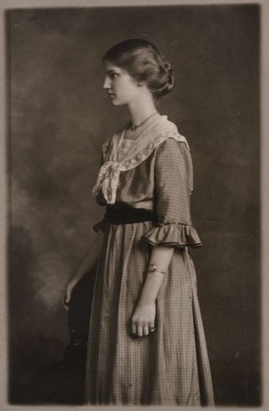 Three-quarter length studio portrait of Jeannette Holt who is standing and facing the left in profile. She is wearing a dress of checkered fabric, with a belt and lace scarf on her shoulders. She is wearing a necklace, bracelet, and ring. 