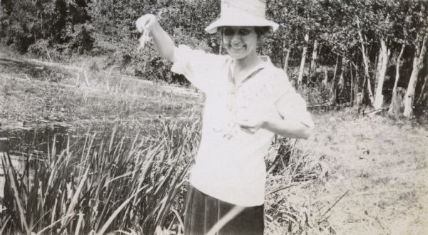 View of a girl holding several frogs by their legs. She presumably caught the frogs in the nearby wetland. Her sun hat is shading part of her face. Archibald Lake with its grasses and tree-lined shore are in the background.