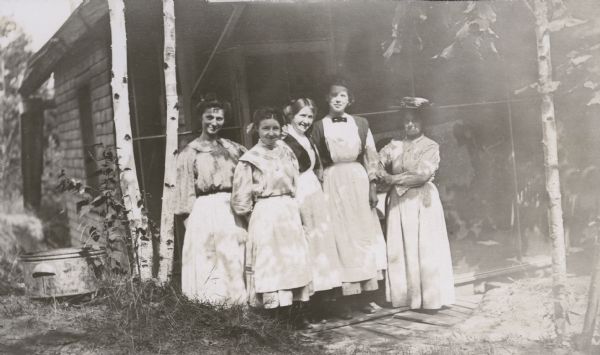 Group portrait of five women, who are household employees, standing together in front of the original Cook’s Cabin, which burned down following a lightning strike, one winter in the 1960s. There is a birch tree and a metal wash tub on the left. 

The caption, which has faded, reads: "Minnie Vendt, Ella Viester, Mrs. -----, Ella Kielel, Katie B------."

