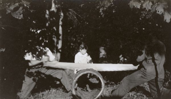 An action shot of two sawyers cutting a hollow hemlock log using a 2-person crosscut saw. They are sawing halfway through the log, to create an angle which will be surfaced with birch branches, offering a shelf for the speaker to put water, eye glasses, sermon text, notes, and/or a Bible. Young David Rumsey, bathed in sun, is watching the sawing. Part of a series captioned: "Building the Pulpit." This pulpit will be used for the Sunday morning Presbyterian worship on the porch at Island Lodge on Archibald Lake.


