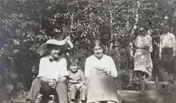 Young David Rumsey is sitting on a log between his father and mother on a sunny day. Parents Wallace and Edna Rumsey are looking at the camera. There is a picnic basket just behind the log on the right. Caption reads: "Wallace and Edna Rumsey and David." Two other people, one of whom may be W.A. Holt, are standing under the trees in the background on the right.

