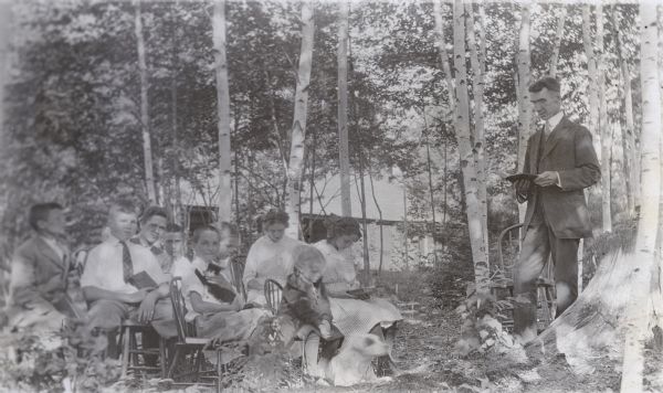 It is a Wednesday in the middle of August. Standing in the woods on the Island, W.A. Holt is reading aloud, possibly scripture or a children's sermon. A group of Holt children and others, sitting on wooden chairs, are listening to W.A. Holt read. In the front row, looking at the camera, Donald Holt is holding a cat. Also in the front row, Jeannette Holt is reading along with her father,and Eleanor Holt is looking down, perhaps at the dog. In the back row, Alfred Holt is wearing glasses and a suit coat. Island Lodge is in the background.