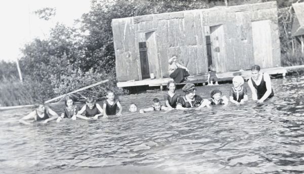 View across water towards a line of Holt children and others who are playing with a long log in Archibald Lake in front of the Bath House. Jeannette Holt is second from the left. Alfred Holt is third from the left. Donald Holt is fourth from the left. Eleanor Holt is seventh from the left. Don Bridgeman, possibly the handyman, is on the far right. A woman and young Jane Trowbridge are sitting on the pier.

