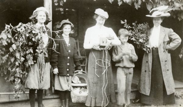 A group of women and children are standing in front of the porch at Island Lodge. From left to right: Jeannette Holt smiling and holding a bundle of vines with grapes; a girl, possibly Margaret or Harriet Stroh, smiling and holding a basket of grapes; Juliet Rumsey Stroh advancing film in a Brownie camera and holding a rope; Alfred Holt; and a woman holding a bunch of greenery. 

  


