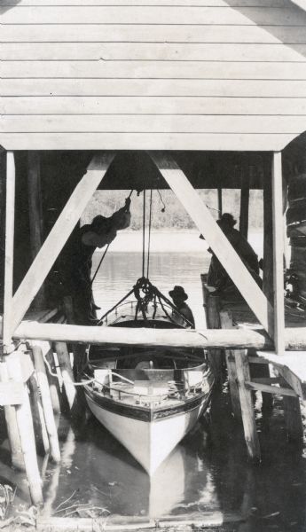 The boathouse is protecting the steam launch, called <i>The Islander</i>, from the elements. The man in the boathouse on the left is pulling a rope. A man is standing in the lake to the right of the launch. Two men on the right are on the pier. Caption reads: "Launch."

