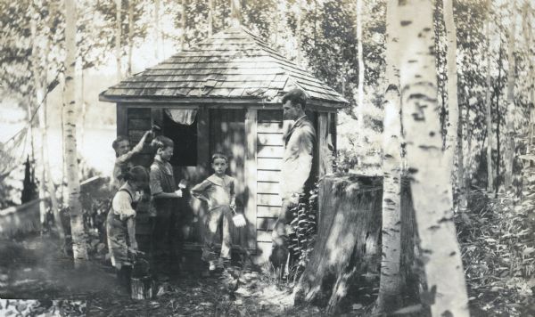W.A. Holt is watching his children paint the cupola which was moved to the Island from the Lakewood school house. From left to right: Jeannette Holt is painting the window frame; Mary Eleanor is dipping her brush in the paint can; Alfred is smiling and holding a paint brush; Donald is looking at the camera. There is a large birch tree to the left of the cupola. Archibald Lake is in the background. Caption reads "Painting Cupola."