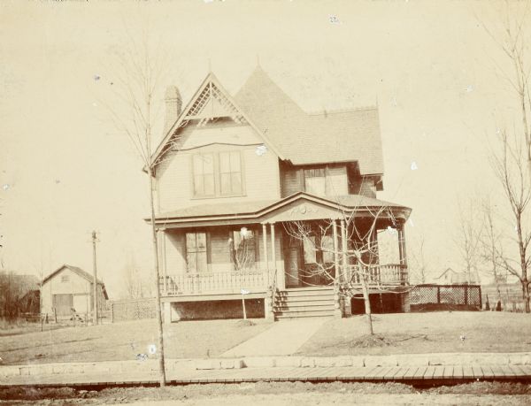 Front view of the two-story Queen Anne style home where Lucy and W.A. Holt lived together starting in 1895. The address is 523 Main Street.

"A front veranda with thin doubled Tuscan columns wraps around the east side of the house and shelters a double doorway. The steeply pitched gables of the roof are trimmed with large geometrically-designed bargeboards and the chimney stack pierces the roofline."  (Source: Wisconsin Historical Society Architecture and History Inventory. Search on Ira Brooks Pendelton House or reference no. 23327.)

The caption of a similar photograph reads "Pine Barren 1895." This is a humorous reference to the family business of timber and logging. A wooden sidewalk parallels the street.