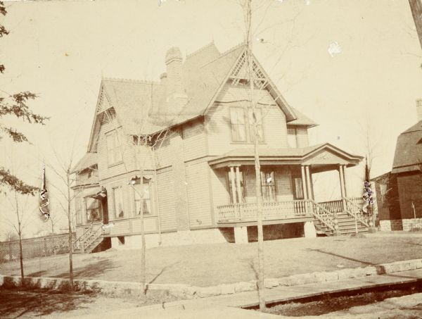 Corner view of the Queen Anne style home where Lucy and W.A. Holt lived together, starting in 1895. The address is 523 W. Main Street. There are no trees on the property.

"A front veranda with thin doubled Tuscan columns wraps around the east side of the house and shelters a double doorway. The steeply pitched gables of the roof are trimmed with large geometrically-designed bargeboards and the chimney stack pierces the roofline."  (Source: Wisconsin Historical Society Architecture and History Inventory. Search on Ira Brooks Pendelton House or reference no. 23327.)

Caption reads: "Pine Barren 1895." This is a humorous reference to the family business of timber and logging. A wooden sidewalk parallels the street.