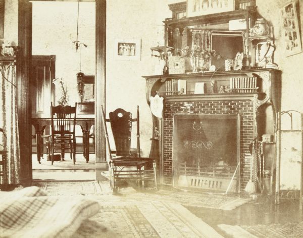 View of library and dining room in the home of Lucy and W.A. Holt. Above the fireplace are books, framed photos, a vase with flowers, and a mirror. Oriental rugs cover most of the library floor. Through the doorway is the dining table, with a potted lily in the center, and dining chairs. A chandelier is hanging from the ceiling in the dining room. Caption reads: "Dining room - Library (W.A.S.)"