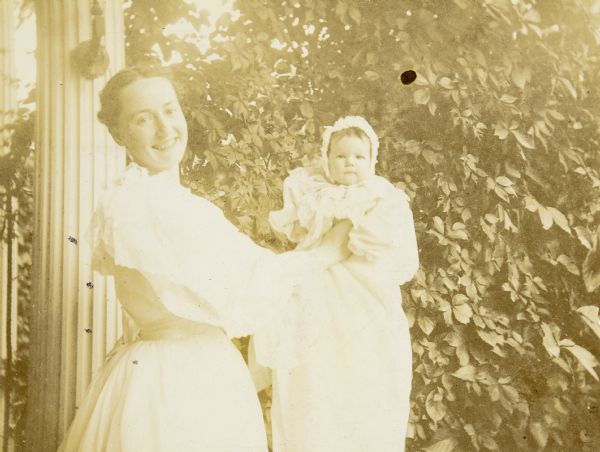 Minnie May Rumsey is smiling and holding her niece Jeannette Holt. The baby is looking at the camera. An architectural column and foliage are in the background. Caption reads: "Aunt May - Jeannette - First Anniversary - July 25, 1896." 