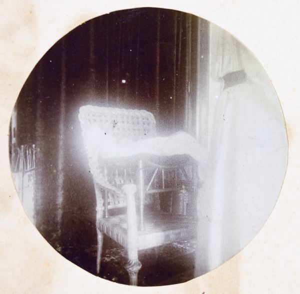 Circular-framed view of Jeannette Holt's bassinet with blanket on a chair at home. A woman wearing a long dress can be seen from the shoulders down standing on the right. A dark-colored curtain is in the background. Caption reads: "Jeannette Rumsey Holt — 4 weeks — Oconto."
