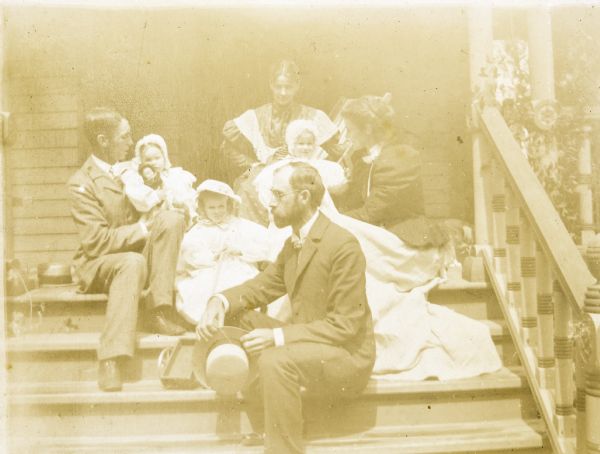 The Holt and Stroh families are sitting together on the front steps of the Holts' home. On the left, W.A. Holt is holding his daughter Jeannette with her doll. One step below Jeannette is her cousin Margaret Stroh. In front of Margaret Stroh is Grant Stroh, seen in profile and holding his hat. Behind Grant Stroh is Harriet Stroh. Behind her is Lucy Rumsey Holt. On the right is Juliet Rumsey Stroh. A toy wagon is sitting on same step as Grant.