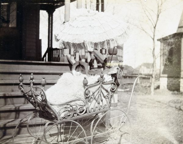 Jeannette Holt is sitting in her wicker baby carriage which is parked on the sidewalk at the base of the porch stairs at home. An umbrella, attached to this ornate perambulator, is shading Jeannette's eyes from the sun. She is wearing a hat, and is covered with a big fuzzy blanket.



