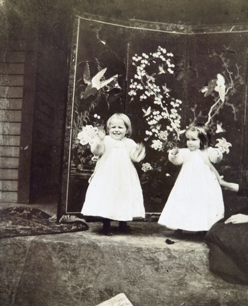 Cousins Harriet Stroh (L) and Jeannette Holt (R) are standing outside on a carpet in front of a lacquer screen decorated with flying birds. On the right is the lap of Juliet Rumsey Stroh (head out of frame) who is kneeling and supporting Jeannette Holt with her right hand. Caption reads: "How do the birdies fly? Jeannette and Harriet Stroh."