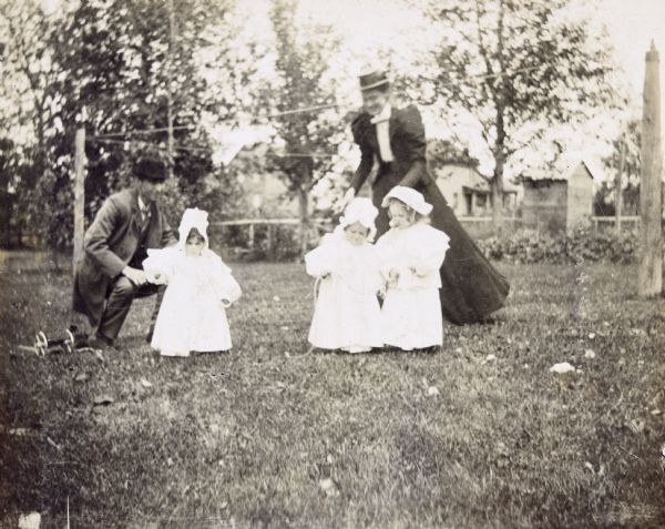 Cousins Jeannette Holt (left), Harriet Stroh (center), and Margaret (right) Stroh are playing in the backyard of the Holt home. W.A. Holt is crouching down near daughter Jeannette. Juliet Stroh is standing and watching her girls as they are playing with a rope. On the far left is a toy wagon on the lawn, next to W.A. Holt. Caption reads: "Jeannette, Harriet, and Margaret."