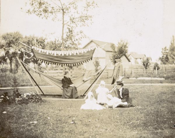 Cousins Jeannette Holt (left) and either Harriet or Margaret Stroh (right) are playing in the backyard under the supervision of grandmother Mary Mathilda Axtell Rumsey who is sitting on the hammock with canopy. Juliet Stroh is sitting on the lawn with the children. W.A. Holt is standing and looking at the camera. In the yard are flowers and trees, and a barn and houses are in the far background.