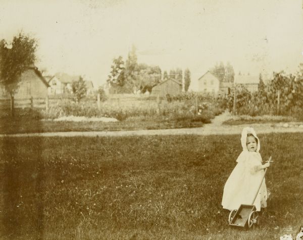 Jeannette Holt is pulling a toy wooden wagon across the lawn in her backyard while looking over her shoulder back towards the camera. In the far background is a fence, what may be a garden, and buildings.