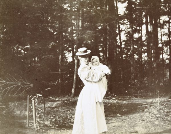 Outdoor portrait of Miss Doud holding baby Alfred Holt in her arms while standing in front of tall trees. Caption reads: "A whooping cough baby — Miss Doud and Alfred."