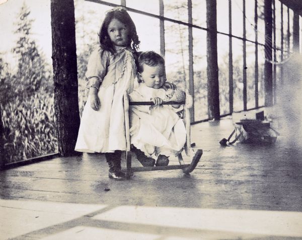 On the screened porch at Island Lodge, baby Alfred Holt is sitting in an infant-sized wooden rocking chair with sister Jeannette standing at his side. She has her arm around Alfred's shoulder. There is a basket on the porch floor in the background, and a hammock is hanging from a post. There are trees in the background. Caption reads: "Jeannette and Alfred."