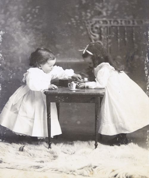 Studio portrait in front of a painted backdrop of Alfred (left) and Jeannette (right) Holt having a tea party. Both children are wearing dresses. A miniature coffee pot, a tiny cup and other items are on the table. There is an animal skin rug on the floor in the foreground. A croquet ball is on the rug, in front of Alfred. Caption reads: "Jeannette 3 years - Alfred 2.5 years."