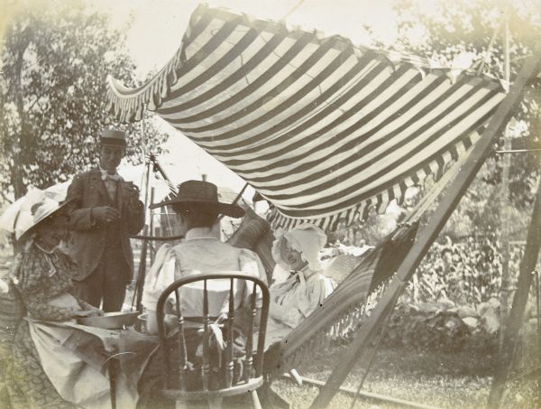 Shaded by a striped canopy, three women are sitting together and smiling in the Holt family backyard. Wearing aprons, the woman on the left and the woman with her back to the camera each have a metal pan and some newspaper pages on their laps. Standing by the group of women, W.A. Holt is holding some food in his left hand.

