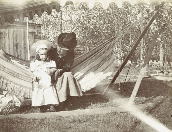 Looking at the camera, Jeannette Holt is standing next to her aunt May in the backyard of the Holt family home. Minnie May Rumsey is sitting on the hammock and looking down at Jeannette. There is a basket under the hammock on the left. There is a canopy over the hammock on the upper left. Caption reads: "Jeannette and Aunt May, May 8, 1899."